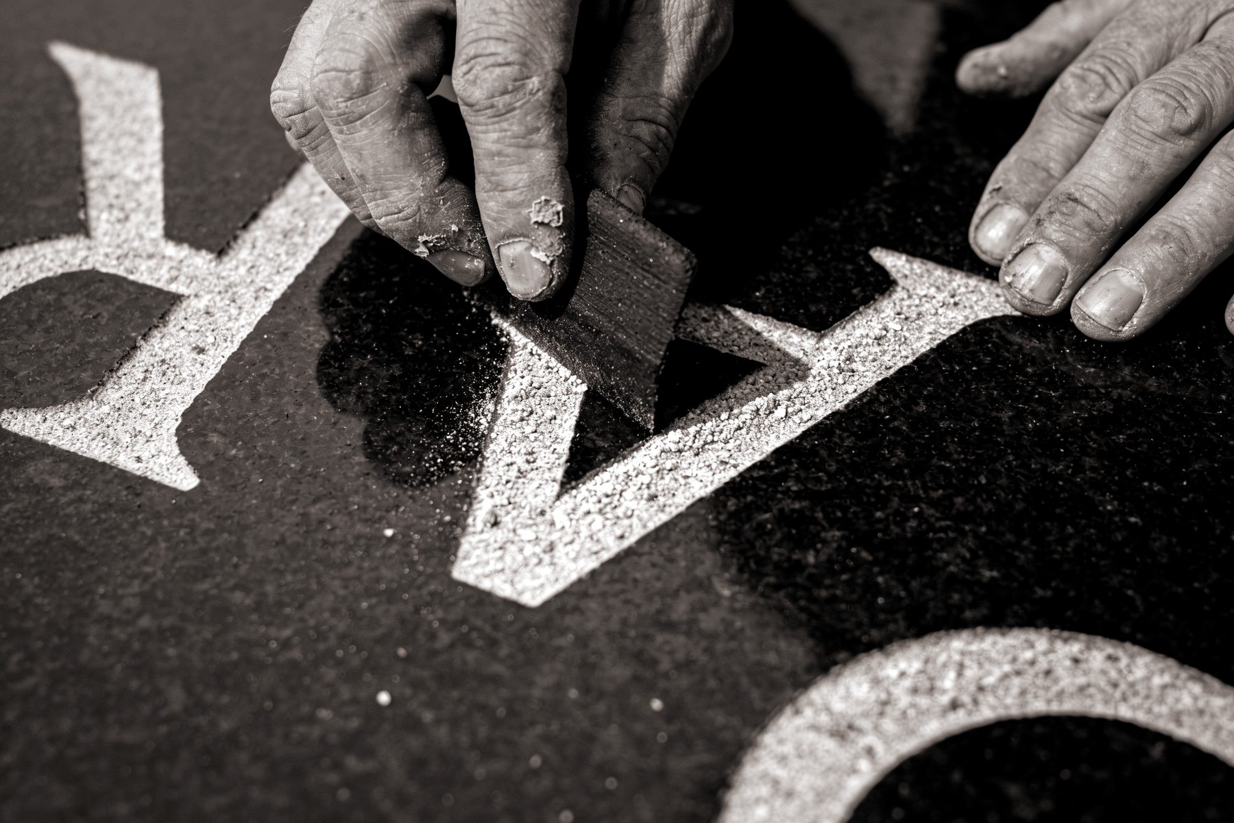 Two weathered hands working a small rubber block across freshly sandblasted letter strokes cut into a dark polished granite marker, pale granite dust still lying in the grooves.